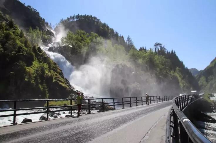 Br&uuml;cke mit Metallgel&auml;nder rechts, gro&szlig;er Wasserfall links mit aufsteigendem Wasserspray, bewaldete Felsen und blauer Himmel, eine Person steht am Gel&auml;nder