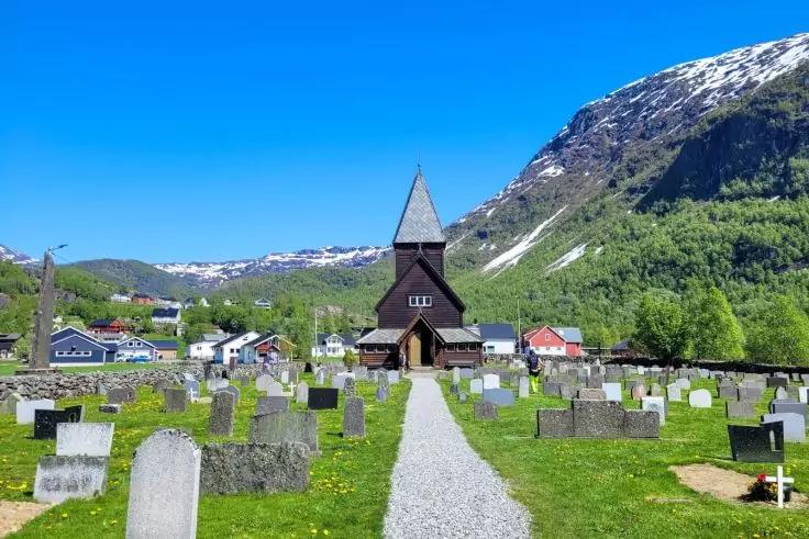 Holzkirche mit spitzem Turm, vor ihr ein Kiesweg, umgeben von Grabsteinen auf gr&uuml;nem Rasen, dahinter H&auml;user, Berge mit Schneefeldern und blauer Himmel