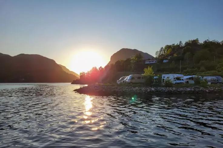 Sonne knapp &uuml;ber dem Horizont zwischen Bergen, Wasser im Vordergrund mit Sonnenreflex, rechts Ufer mit geparkten Wohnmobilen und B&auml;umen