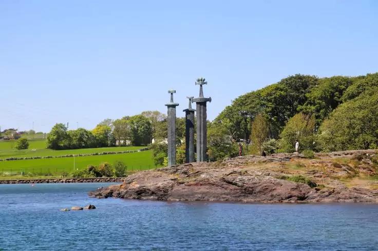 Drei hohe dunkle S&auml;ulen mit kreuz&auml;hnlichen Aufs&auml;tzen auf felsiger K&uuml;ste, blaues Meer im Vordergrund, gr&uuml;ne Wiesen und B&auml;ume im Hintergrund, heller blauer Himmel