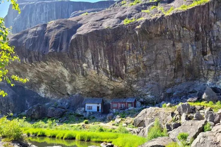 Zwei kleine H&auml;user unter einem hohen Fels&uuml;berhang, davor Wasserfl&auml;che, Gras und Felsen