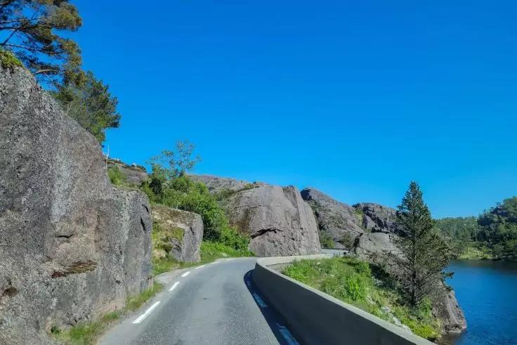 Schmale asphaltierte Stra&szlig;e mit Leitplanke rechts, die zwischen felsigen Klippen links und einem See rechts kurvt; blauer Himmel, vereinzelte B&auml;ume und Gras