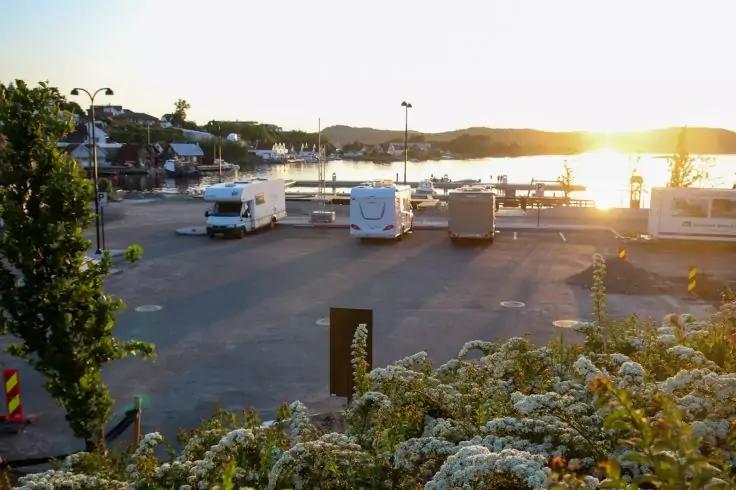 Parkplatz am Wasser mit mehreren Wohnmobilen, einem Lkw, einem Bootssteg und bl&uuml;henden Str&auml;uchern im Vordergrund; Sonne am Horizont