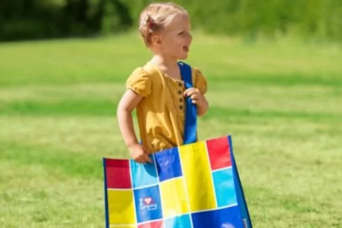 Kleines M&auml;dchen steht auf Rasen und h&auml;lt eine gro&szlig;e, bunte Einkaufstasche mit blauem Tragegurt, Blick nach rechts