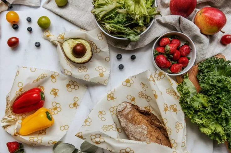 Brotlaib, halbierte Avocado mit Kern, Sch&uuml;ssel mit Erdbeeren, rote und gelbe Paprika, verstreute Blaubeeren und kleine Tomaten, gr&uuml;ne Limetten, Kopf- und Blattgem&uuml;se und rote &Auml;pfel auf einem gemusterten Tuch