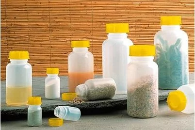 Mehrere durchscheinende wei&szlig;e Kunststoffflaschen mit gelben Verschl&uuml;ssen, teils mit farbigen Pulvern und Fl&uuml;ssigkeiten gef&uuml;llt, auf einer Steinplatte vor einer Bambusmatte