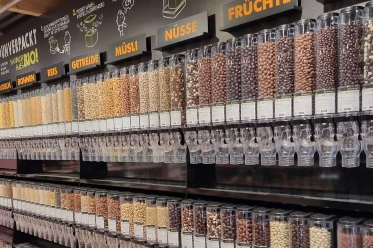 Regal mit mehreren Reihen transparenter Spender, gef&uuml;llt mit K&ouml;rnern, N&uuml;ssen und M&uuml;sli; oben Schilder mit Kategorien