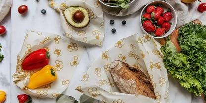 Mehrere T&uuml;cher mit gelbem Blumenmuster auf wei&szlig;er Oberfl&auml;che, dazu ein Laib Brot, halbierte Avocado, rote und gelbe Paprika, eine Schale mit Erdbeeren, Salatbl&auml;tter, Blaubeeren und einzelne Kirschtomaten