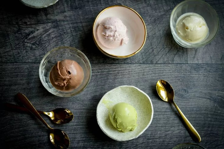 Mehrere kleine Schalen mit je einer Eiskugel in rosa, wei&szlig;, braun und hellgr&uuml;n auf dunklem Holztisch, goldene L&ouml;ffel daneben