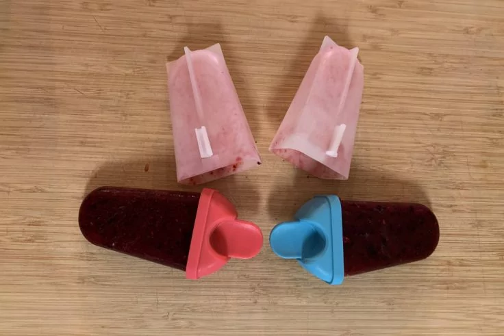 Vier Eis am Stiel auf einem Holzbrett: oben zwei helle, halbierte Eisformen mit wei&szlig;en Stielen; unten zwei dunkle ganze Eis mit roten und blauen Griffen