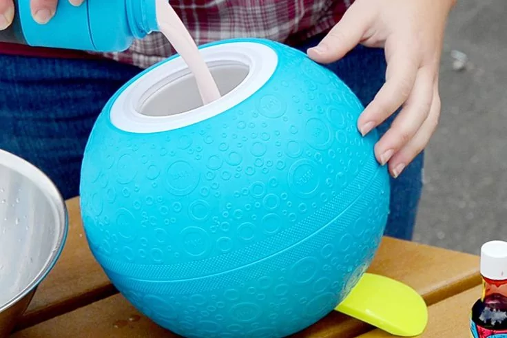 Zwei H&auml;nde halten eine blaue, strukturierte Kunststoffkugel mit gro&szlig;er &Ouml;ffnung; wei&szlig;e Fl&uuml;ssigkeit wird aus einem Beh&auml;lter hineingegossen; auf dem Holztisch daneben liegen ein gelber L&ouml;ffel und eine kleine Flasche mit roter Kappe