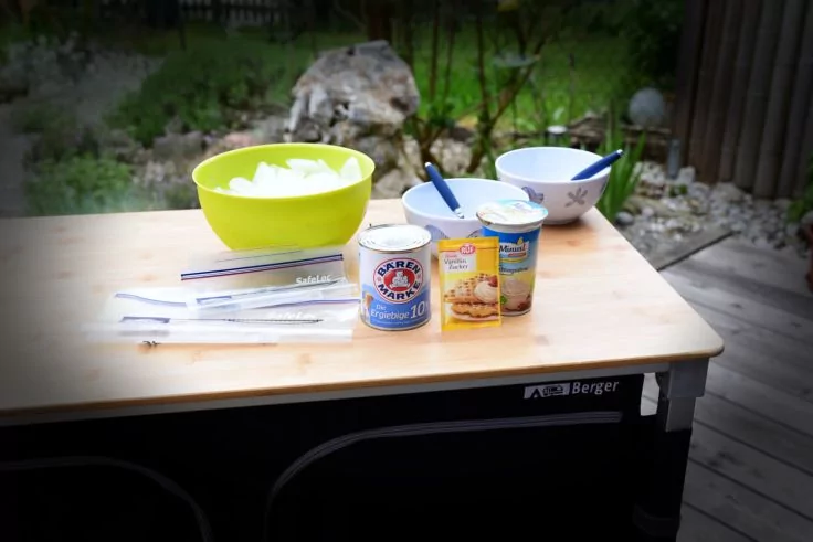 Holztisch auf Terrasse mit gr&uuml;ner Kunststoffsch&uuml;ssel mit Eisw&uuml;rfeln, zwei Keramiksch&uuml;sseln mit L&ouml;ffeln, zwei bedruckte Becher und mehrere verpackte Plastikl&ouml;ffel