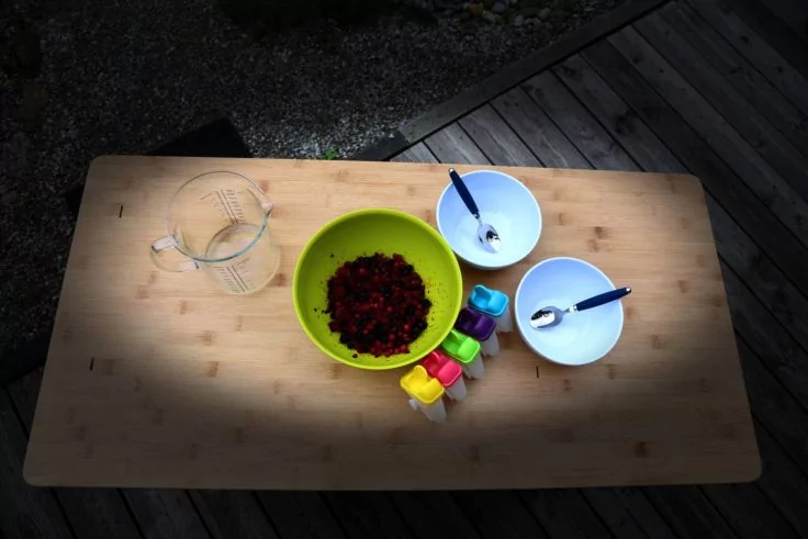 Holztisch mit transparentem Messbecher links, gr&uuml;ne Sch&uuml;ssel mit kleinen roten St&uuml;cken in der Mitte, Reihe bunter Messbecher vorn, zwei wei&szlig;e Schalen mit L&ouml;ffeln rechts, Holzdeck und Kies im Hintergrund, kreisf&ouml;rmiger Lichtfleck auf dem Tisch