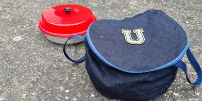 Dunkelblaue runde Stofftasche mit hellblauem Rei&szlig;verschluss und wei&szlig;em 'U'-Aufn&auml;her, daneben ein roter runder Beh&auml;lter mit schwarzem Knopf, beide auf grauem Asphalt
