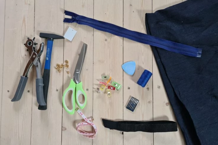 Heller Holzboden mit Rundlochzange, kleinem Hammer, grüner Schere, Reißverschluss, dunkelblauem Stoff, Maßband, Stecknadeln, Nähnadeln in Verpackung, goldenen Nieten, bunten Stoffklammern, blauem Stoffmarker und schwarzem Stoffstreifen