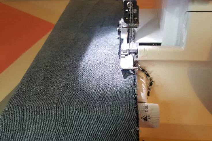 Nahaufnahme einer Nähmaschine mit Nähfuß, grauem Stoff unter dem Fuß und einer genähten Stoffkante