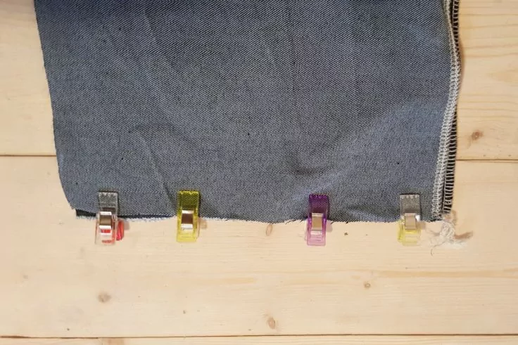 Grauer Stoff mit Reißverschluss am rechten Rand, am unteren Saum mit vier bunten Kunststoffklammern befestigt, auf hellem Holzbrett