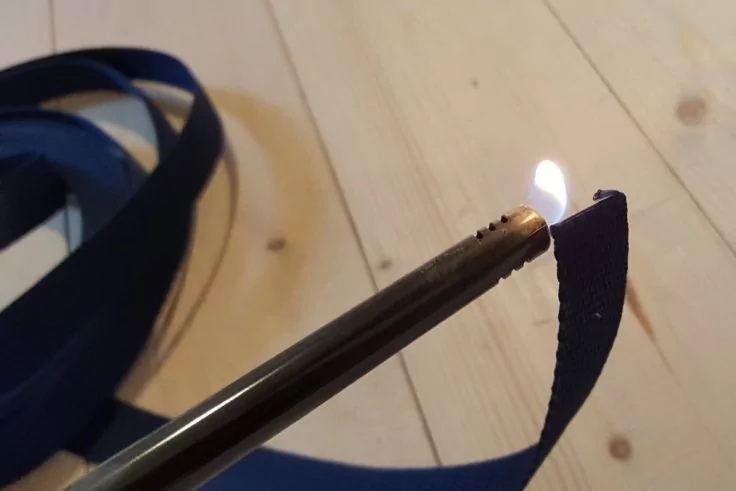 Langer Metallanzünder mit kleiner Flamme an einem blauen Stoffband; aufgewickeltes blaues Band und heller Holzboden im Hintergrund