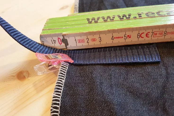 Gefalteter grüner Zollstock mit Zentimeter-Skala, dunkler Jeansstoff mit Overlockkante, blaues Webband, rosa durchsichtige Klammer, helle Holzfläche im Hintergrund