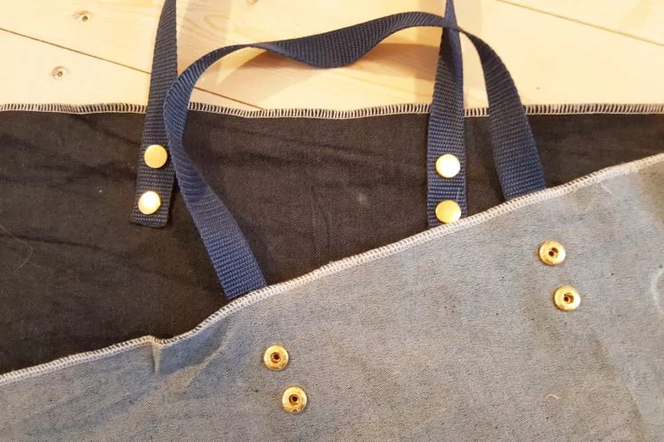 Graue Canvastasche mit sichtbarer Reißverschlusskante, mehreren goldfarbenen Druckknöpfen und dunkelblauen Tragegurten auf schwarzem Stoff