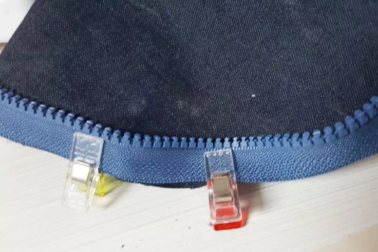 Dunkelblauer Stoff mit blauem Kunststoffreißverschluss und zwei transparenten Klammern mit gelbem und rotem Fuß auf weißer Fläche