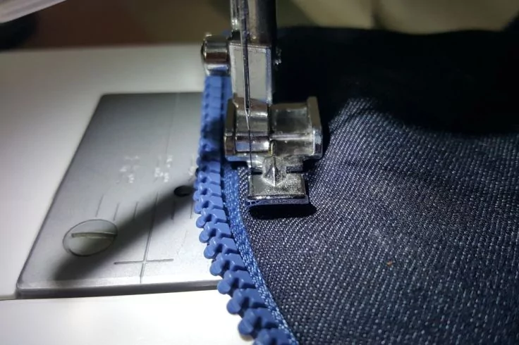 Nähmaschinenfuß und Nadel über einem blauen Kunststoffreißverschluss auf dunkelblauem Stoff