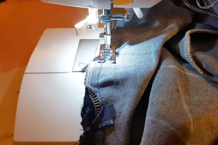 Nähmaschine mit Nadel und Nähfuß, die an einer dunkelblauen Jeans näht; weißes Gehäuse, helle Beleuchtung und orangefarbener Hintergrund sichtbar