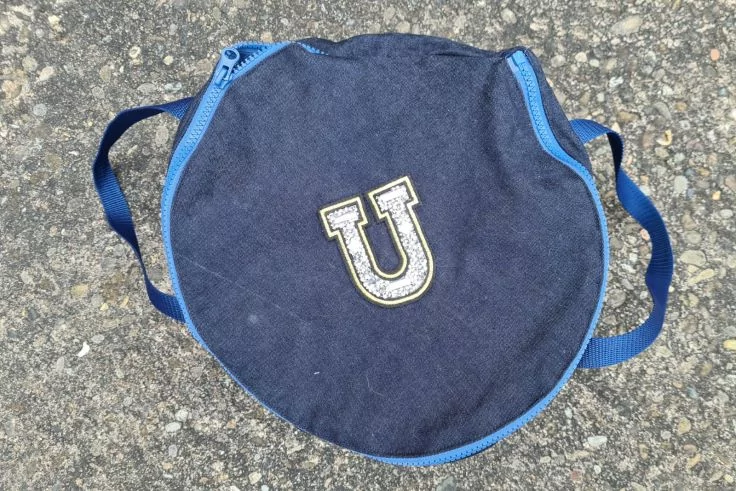 Runder dunkelblauer Stoffkreis mit blauem Rand und zwei Trägern, in der Mitte ein weiß-gelber Buchstabe U, liegend auf Asphalt