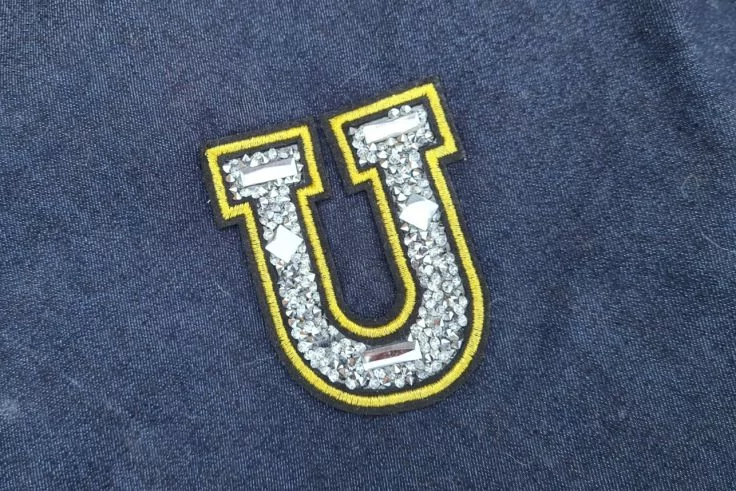 Auf dunkelblauem Stoff ein aufgenähter Buchstabe ‚U‘ aus silbernen Pailletten mit gelbem Rand