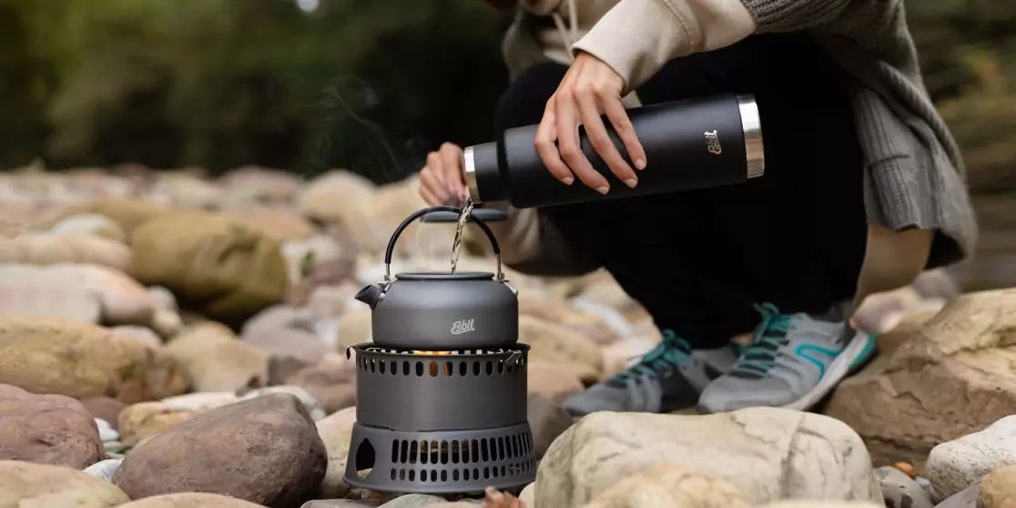 Teilweise sichtbare Person kniet auf runden Steinen und gie&szlig;t aus einer schwarzen Thermoskanne in ein graues tragbares Kochger&auml;t