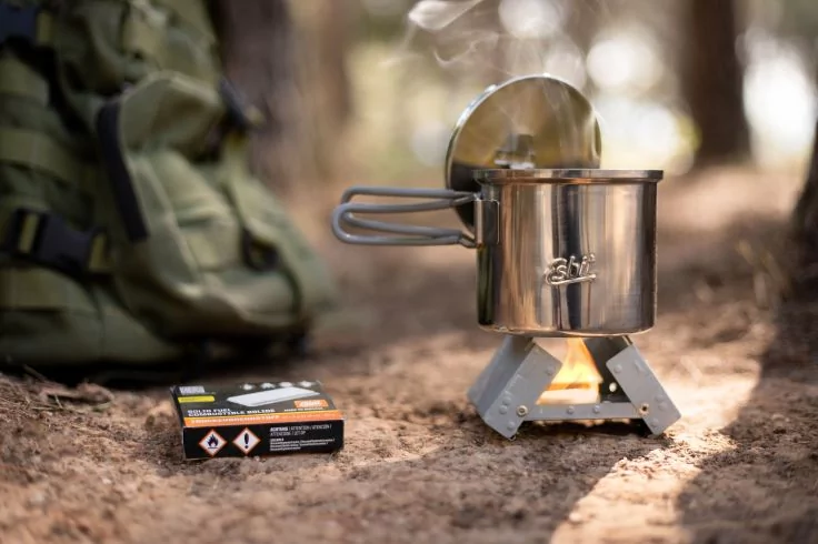 Edelstahl-Kochtopf mit Deckel auf kleinem Campingkocher mit sichtbarer Flamme und Rauch, daneben eine Packung Brennstoff und ein Rucksack auf waldbedecktem Boden