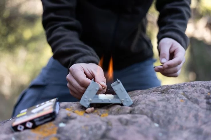 H&auml;nde z&uuml;nden eine kleine faltbare Metallvorrichtung mit sichtbarer Flamme auf einem Felsen; eine kleine Schachtel liegt links