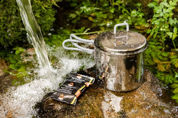 Edelstahl-Topf mit Deckel auf nassem Felsen neben flie&szlig;endem Wasser; drei schwarze Einzelpackungen liegen daneben; gr&uuml;ne Pflanzen und Moos im Hintergrund