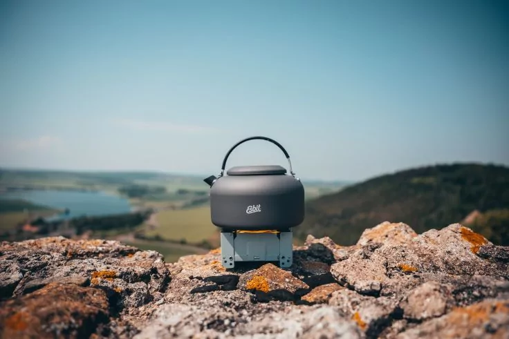 Kleines graues Ger&auml;t mit Henkel auf felsigem Untergrund; im Hintergrund See, gr&uuml;ne H&uuml;gel und blauer Himmel