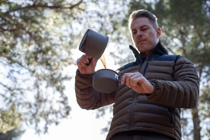 Ein Mann gie&szlig;t Fl&uuml;ssigkeit von einem Metallkessel in einen kleinen Metallbecher, vor B&auml;umen; er tr&auml;gt eine gesteppte Jacke