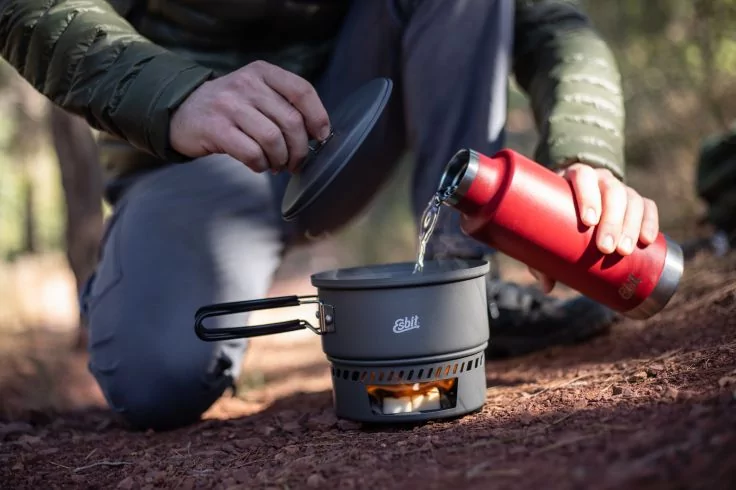 H&auml;nde gie&szlig;en Wasser aus roter Thermosflasche in grauen Campingtopf auf kleinem Kocher, brauner Boden, verschwommene B&auml;ume im Hintergrund