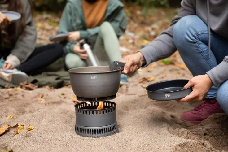 Person h&auml;lt Topf und Deckel &uuml;ber einem kleinen Campingkocher mit sichtbarer Flamme; zwei Personen sitzen im Hintergrund auf dem Sand, Bl&auml;tter liegen auf dem Boden