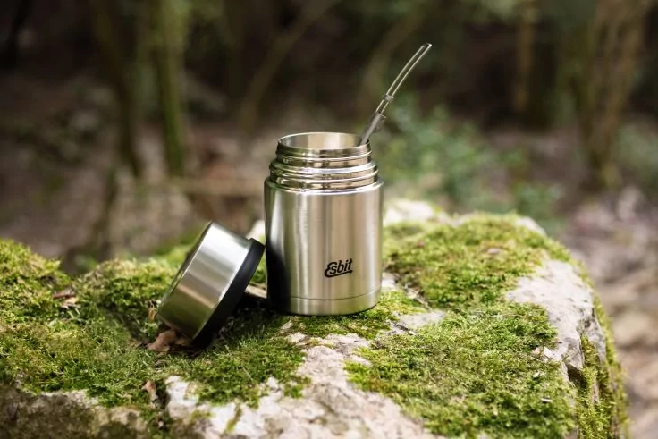 Edelstahl-Thermobeh&auml;lter mit abgenommenem Deckel daneben und Metallstrohhalm, auf moosbedecktem Felsen, unscharfer Waldhintergrund