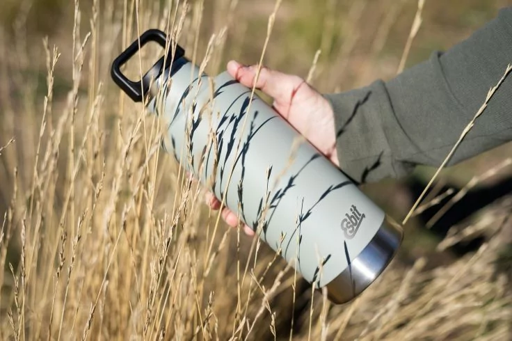 Hand h&auml;lt hellgraue Trinkflasche mit schwarzem Karabiner; hohes trockenes Gras im Vordergrund; &Auml;rmel einer dunkelgr&uuml;nen Jacke sichtbar