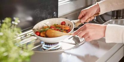 Zwei H&auml;nde halten eine wei&szlig;e Pfanne mit Kirschtomaten, Kr&auml;utern und einem Holzl&ouml;ffel &uuml;ber einer blauen Gasflamme auf einem Herd