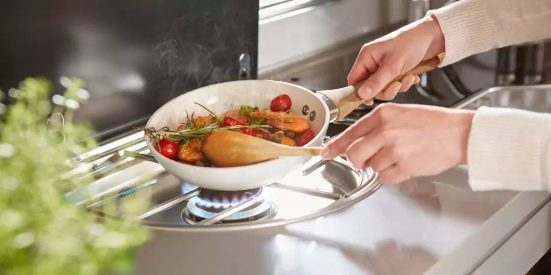 Zwei H&auml;nde halten eine wei&szlig;e Pfanne mit Kirschtomaten, Kr&auml;utern und einem Holzl&ouml;ffel &uuml;ber einer blauen Gasflamme auf einem Herd