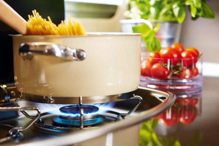 Topf mit herausragenden Spaghetti &uuml;ber einer blauen Gasflamme, im Hintergrund Schale mit Kirschtomaten und Basilikum