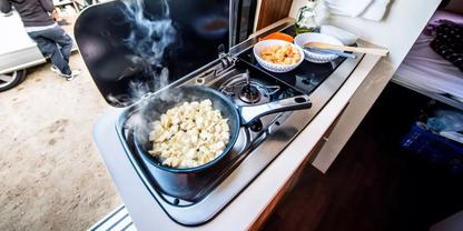 Zweiflammiger Gasherd in einer K&uuml;chenzeile: Bratpfanne mit dampfenden Teigst&uuml;cken, daneben Sch&uuml;ssel mit orangefarbener Beilage und Essst&auml;bchen
