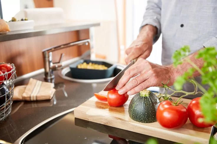 H&auml;nde schneiden eine Tomate auf einem Holzbrett; daneben liegen weitere Tomaten und eine runde Zucchini, im Hintergrund Pfanne und Sp&uuml;le