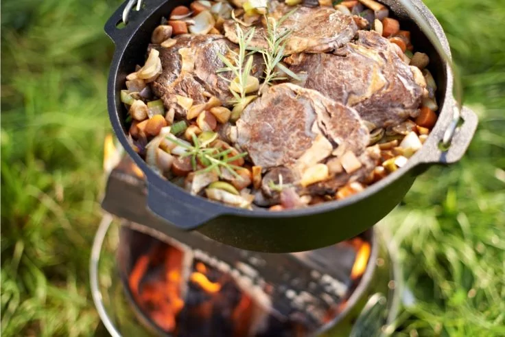 Gusseisentopf mit drei Fleischst&uuml;cken auf einer Schicht aus Bohnen und Gem&uuml;se, mit Rosmarinzweig, auf einem St&auml;nder &uuml;ber offenem Feuer, Gras im Hintergrund