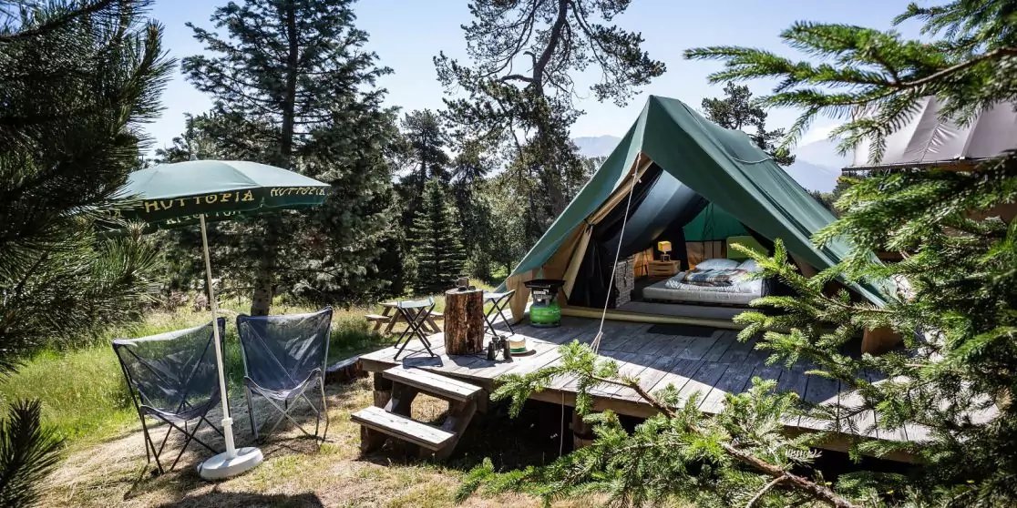 Gr&uuml;nes Zelt auf Holzplattform im Wald, zwei Campingst&uuml;hle und Sonnenschirm neben kleinem Holztisch, B&auml;ume im Hintergrund