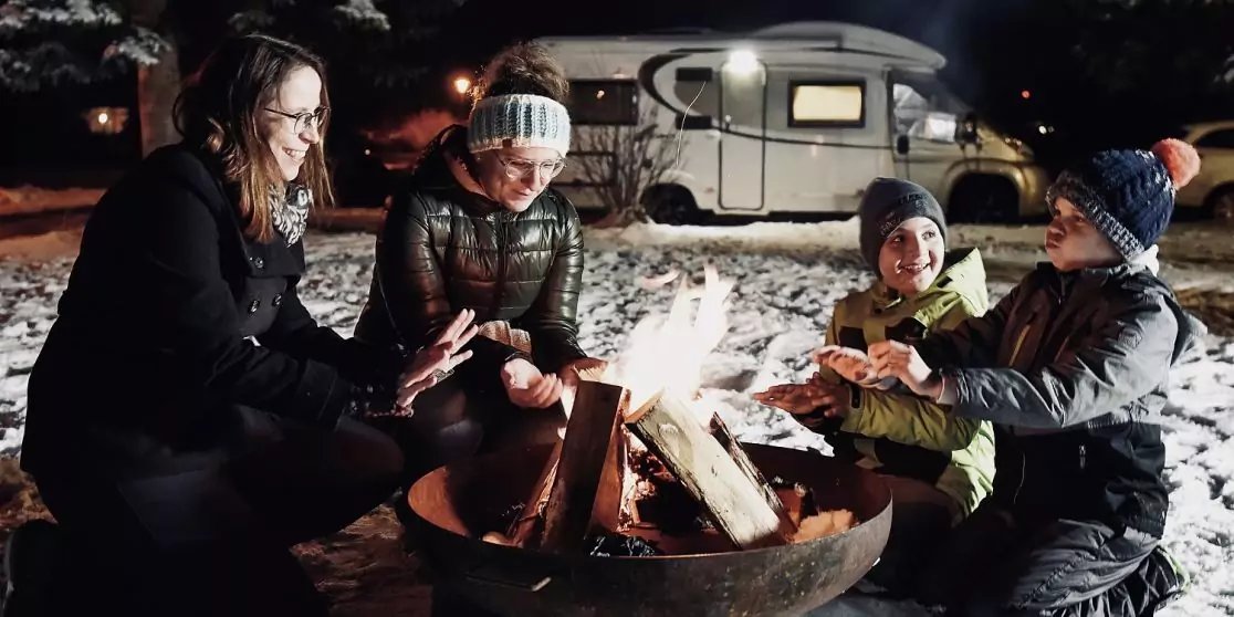 Vier Personen sitzen um ein Lagerfeuer im Schnee; einige halten lange St&ouml;cke &uuml;ber die Flammen; im Hintergrund steht ein beleuchtetes Wohnmobil