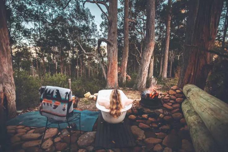 Person mit langen Haaren sitzt in freistehender Badewanne im Wald, im Vordergrund Steine und Baumst&auml;mme, links Campingstuhl mit Tasche