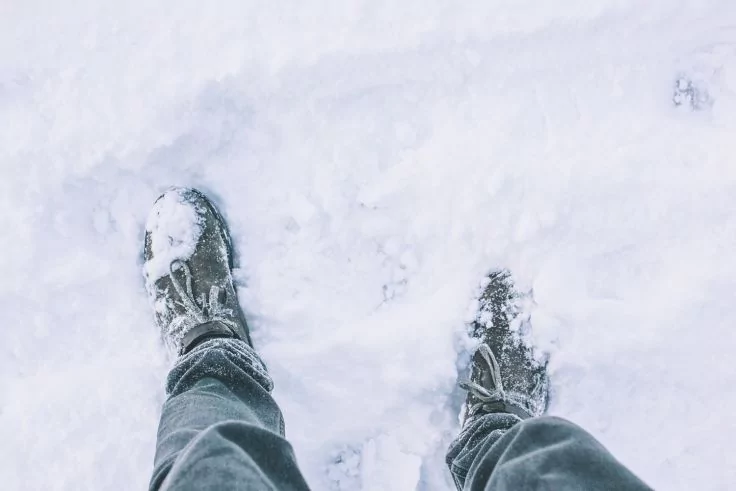 Blick nach unten auf zwei mit Schnee bedeckte Stiefel und Jeans auf verschneitem Boden