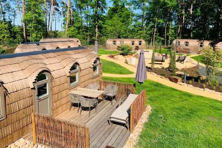 Halbrunde Holzh&auml;user mit Terrassen, Tischen, St&uuml;hlen und Sonnenschirm, Rasenfl&auml;chen, Wege und B&auml;ume im Hintergrund